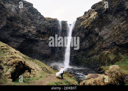 Der Bräutigam umkreist die Braut in seinen Armen nahe dem Wasserfall, es schneit. Reiseziel Island Hochzeit, in der Nähe des Kvernufoss Wasserfals Stockfoto