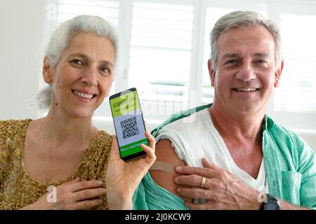 Lächelndes kaukasisches Seniorenpaar zeigt Smartphone mit covid 19 Impfpass auf dem Bildschirm Stockfoto