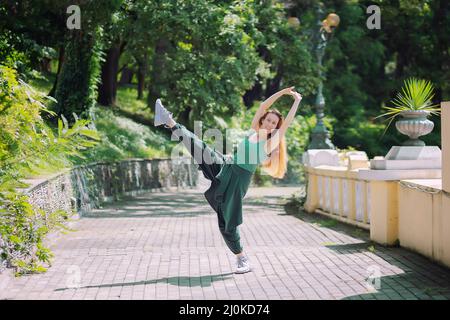 Lächelnd in einer Pose, wobei ihr Bein und ihre Hände hoch in einer Gasse im Park gehalten wurden. Tanzpose Stockfoto