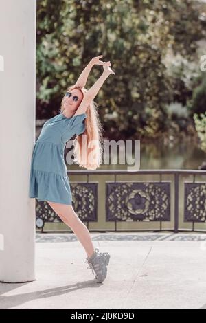 Ein lächelndes Mädchen in einem kurzen Kleid posiert auf einer Säule, die auf ihren Zehen steht und ihre Hände gegen die Rückenlehne hebt Stockfoto