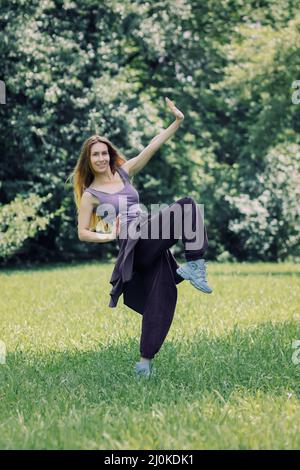 Lächelndes Mädchen im fliederfarbenen Hip-Hop-Outfit in einer Tanzpose auf dem Rasen im Park Stockfoto