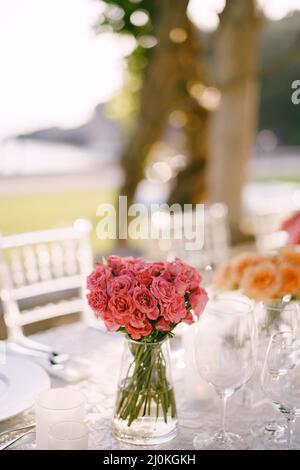 Hochzeitsessen Tischempfang. Ein Bouquet aus kleinen Pfingstrosen rosa Rosen in einer transparenten Glasvase auf einem Tisch in der Sonne, mit dem Stockfoto