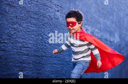 Laufen Sie ab, um den größten Spaß zu haben. Aufnahme eines entzückenden kleinen Jungen, der ein Superhelden-Kostüm trägt, während er im Freien spielt. Stockfoto