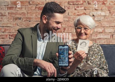 kaukasische Verwandte Großmutter und erwachsener Enkel vergleichen alte gedruckte Fotos mit neuer moderner Technologie. Hochwertige Fotos Stockfoto