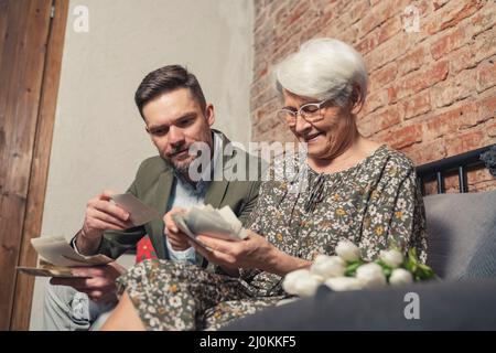 Die ältere Rentnerin zeigt Bilder ihres tausendjährigen bärtigen Sohnes aus ihrer Jugend und Vergangenheit. Hochwertige Fotos Stockfoto