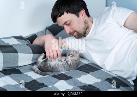 Thema ist Liebe, Pflege und Schutz von Haustieren. Mann, der sich im Bett mit der babygrauen schottischen Katze sonnt. Männliche Umarmungen und Striche Katze A Stockfoto