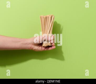 Weibliche Hand hält Einweg-Holzstäbchen zum Rühren von heißen Getränken auf grünem Hintergrund. Kaffee- und Teelöffel Stockfoto