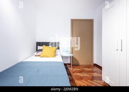 Zimmer mit weißem Kleiderschrank, kleinem Bett, runder blauer Nachttisch und Mahagoni-Fischgrätparkett Stockfoto