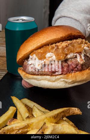 Eine Hand, die ein Stück Rindfleisch-Burger mit viel Frischkäse und karamellisierten Zwiebeln neben einem Stapel hausgemachter Pommes frites hält Stockfoto