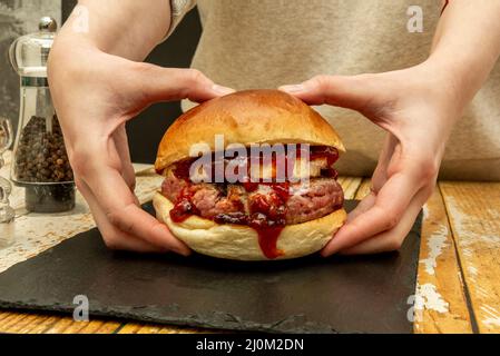 Köstlicher Rinderburger mit Ziegenkäse und viel würziger Tomatenmarmelade, die in zwei Händen auf einem schwarzen Schieferteller und Pfefferkörnern gehalten wird Stockfoto