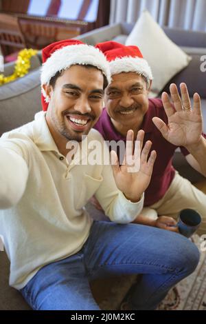 Porträt eines glücklichen erwachsenen Sohnes und älteren Vaters aus Birazialen in Weihnachtsmützen, der einen Weihnachtsvideoanruf führt Stockfoto