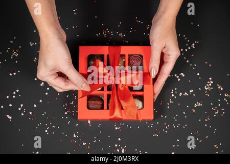 Verschiedene handgemachte Schokoladentrüffel-Bonbons in einer roten Schachtel auf dunklem Hintergrund in weiblichen Händen. Stockfoto