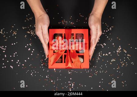 Verschiedene handgemachte Schokoladentrüffel-Bonbons in einer roten Schachtel auf dunklem Hintergrund in weiblichen Händen. Stockfoto