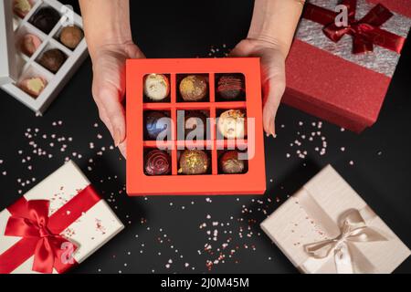 Verschiedene handgemachte Schokoladentrüffel-Bonbons in einer roten Schachtel auf dunklem Hintergrund in weiblichen Händen. Stockfoto