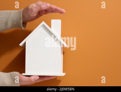 Weibliche Hand halten ein weißes Miniatur-Holzhaus auf einem braunen Hintergrund. Immobilienversicherungskonzept, Umweltschutz, Stockfoto