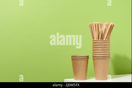 Braune Pappbecher und hölzerne Rührstäbe auf einem weißen Tisch, grüner Hintergrund. Umweltfreundliches Geschirr Stockfoto