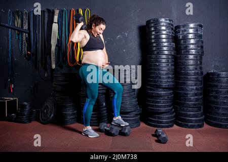 Starke Schwangerin posiert mit Kurzhanteln in den Händen vor dem Hintergrund von Sportgeräten im Fitnessstudio. Workouts und Übungen f Stockfoto