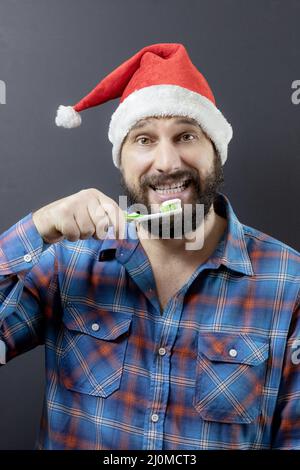 Ein fröhlicher bärtiger Mann in einem Weihnachtsmütze mit einer Zahnbürste. Gefälschter weihnachtsmann. Vorbereitung auf Weihnachten Stockfoto