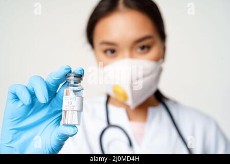 Impfungen und Covid-19-Präventionskonzept. Nahaufnahme Porträt des asiatischen Arztes in Atemschutzmaske zeigt Flasche mit Coronavirus Vacci Stockfoto