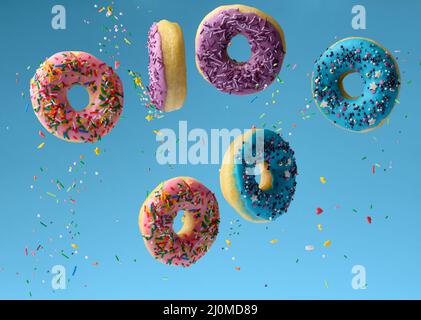 Süße bunte Donuts schweben auf blauem Hintergrund, Zuckerstreuseln fliegen umher Stockfoto