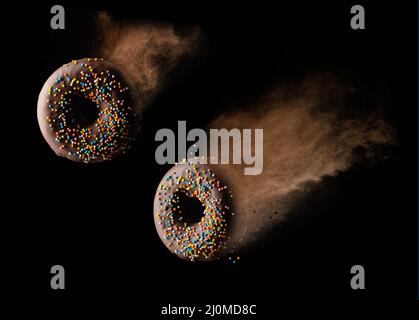 In einer Wolke aus braunem Kakao auf schwarzem Hintergrund schweben runde Schokoladendonuts mit bunten Zuckerstreuseln Stockfoto