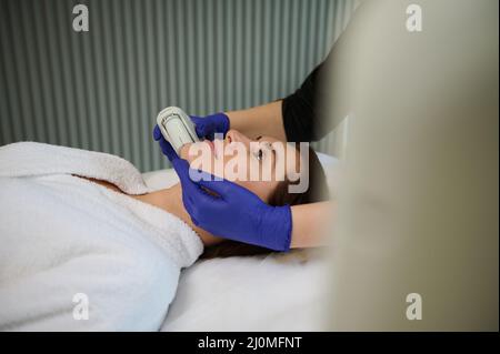 Seitenansicht einer charmanten Frau, die während Schönheitsbehandlungen auf ihrem Gesicht mit einem modernen Ultraschall- oder Lasergerät in einem Verzeiher auf einer Massageliege liegt Stockfoto