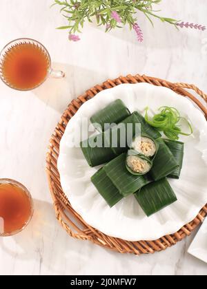 Draufsicht Lemper Ayam, gedämpfter klebrigem Reis gefüllt mit geschreddertem Huhn, eingewickelt in Bananenblatt auf weißem Tisch. Serviert mit Tee Stockfoto