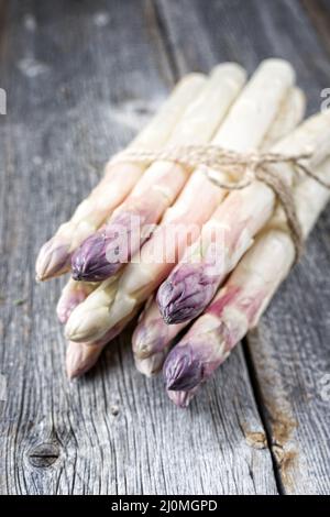 Roher weißer Spargel mit violettem Kopf als Bund auf einem alten rustikalen Holzbrett als Nahaufnahme Stockfoto