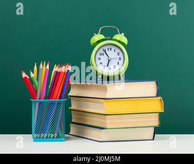 Stapel von Büchern, einem runden Wecker und bunten Bleistiften auf dem Hintergrund einer leeren grünen Kreidetafel Stockfoto