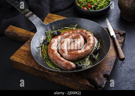 Traditionelle frittierte italienische Salsiccia-Fresko-Fleischwurst mit Rucola und Oliven in einer gusseisernen Pfanne serviert Stockfoto