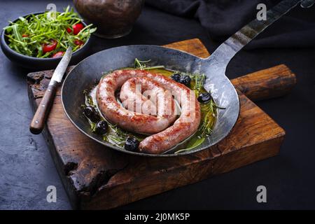 Traditionelle frittierte italienische Salsiccia-Fresko-Fleischwurst mit Rucola und Oliven in einer gusseisernen Pfanne serviert Stockfoto