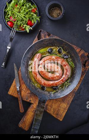 Traditionelle gebratene italienische Salsiccia-Fresko-Fleischwurst, serviert mit Rucola und Oliven als Draufsicht in einer gusseisernen Pfanne Stockfoto
