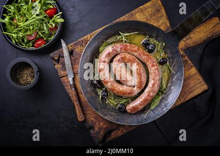 Traditionelle gebratene italienische Salsiccia-Fresko-Fleischwurst, serviert mit Rucola und Oliven als Draufsicht in einer gusseisernen Pfanne Stockfoto