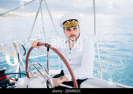 Ein Mann mit Kapitänskappe sitzt am Steuer auf einer Yacht, die mitten im Meer segelt Stockfoto