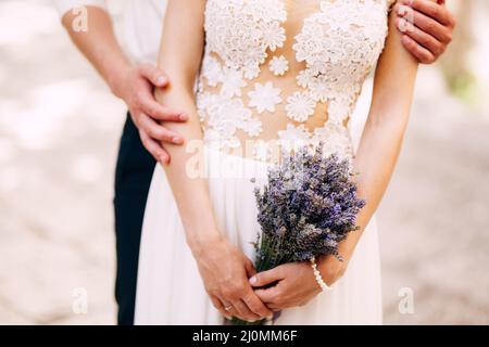 Bräutigam umarmt die Braut von hinten in einem weißen Spitzenkleid mit einem Lavendelstrauß in den Händen. Nahaufnahme Stockfoto