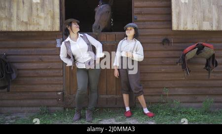 Mutter und Tochter in stilvoller Vintage-Kleidung stehen auf dem Bauernhof in der Nähe des Stalls und ein Pferd guckt aus dem Tor. Hohe Qualität Stockfoto