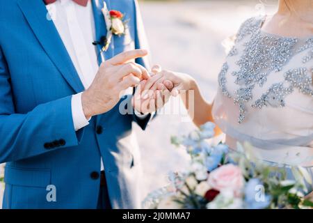 Bräutigam in einem blauen Anzug legt einen Ehering mit einem Blumenstrauß auf den Finger der Braut. Nahaufnahme Stockfoto