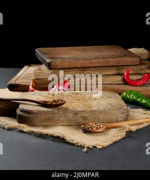 Leere Küche braunes Holzschneidebrett auf schwarzem Tisch, neben Löffeln mit Gewürzen und frischem Chili, schwarzer Hintergrund Stockfoto