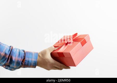 Eine Männerhand in einem karierten Ärmel hält eine rote Schachtel mit einem Geschenk auf weißem Hintergrund. Geschenk für eine Feier Stockfoto