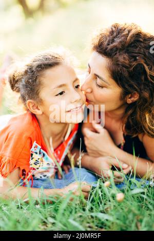 Mama küsst ihre kleine Tochter auf die Wange, während sie auf dem grünen Gras im Park liegt Stockfoto