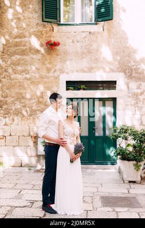 Sibenik, Kroatien - 05.06.17: Der Bräutigam umarmt die Braut von hinten in einem weißen Spitzenkleid mit Lavendelbukett und steht im Innenhof Stockfoto