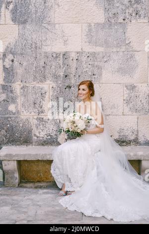 Braut in einem weißen Kleid mit einem Blumenstrauß sitzt auf einer Bank in der Nähe einer Steinmauer Stockfoto