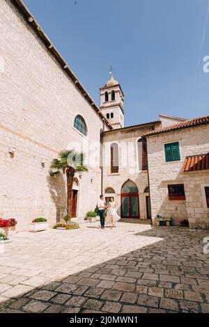 Sibenik, Kroatien - 05.06.17: Bräutigam und Braut in einem weißen Spitzenkleid gehen entlang des Hofes eines alten Gebäudes vorbei an Kübeln mit gr Stockfoto