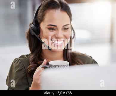 Einen weiteren erfolgreichen Tag durchstehen. Aufnahme eines jungen Callcenter-Agenten, der Kaffee trinkt, während er an einem Computer in einem Büro arbeitet. Stockfoto