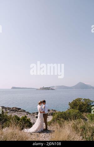 Bräutigam und Braut stehen umarmt am Meer vor der Kulisse der Berge Stockfoto