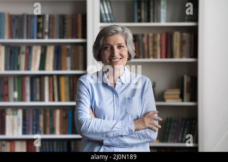 Eine glückliche, gealterte Frau, die mit gekreuzten Armen auf dem Bücherregal steht Stockfoto