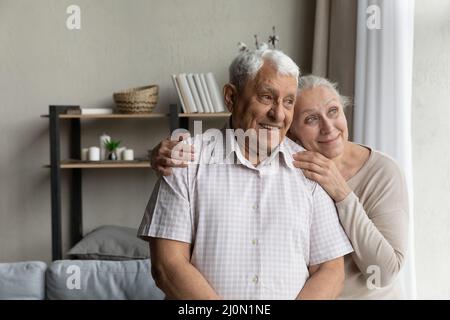 Ein älteres, liebevolles Paar, das im Wohnzimmer steht und aus dem Fenster blickt Stockfoto
