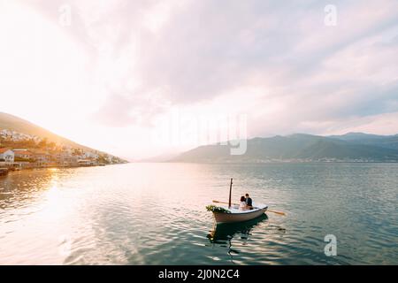 Mann rollt Frau in einem Boot mit Blumen auf dem Wasser auf den Bergen und Sonnenuntergang Stockfoto