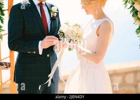 Der Bräutigam in einem schwarzen Karo-Anzug legt einen Ehering in einem weißen Kleid mit einem Blumenstrauß an den Finger der Braut Stockfoto