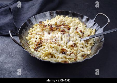 Der traditionelle schwäbische Käsespätzle mit gebratenen Zwiebeln wurde als Nahaufnahme in einer gusseisernen Bratpfanne auf einem rustikalen schwarzen Brett serviert Stockfoto